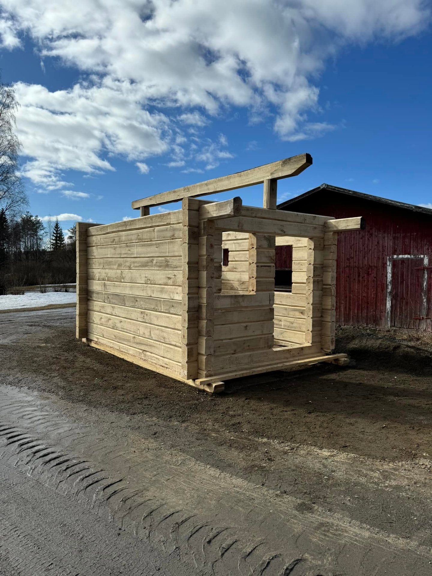 Pienen hirsirakennuksen valmis hirsikehikko veistopaikalla punaisen ladon vierellä.